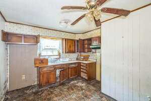 A worn kitchen with damaged wooden cabinets, missing drawers, an old ceiling fan, peeling wallpaper, dirty floor tiles, and a small window with a lace curtain letting in natural light.
