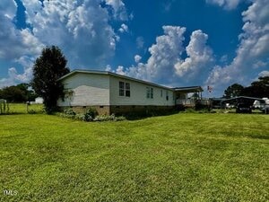 A single-story manufactured home with white siding sits on a large, neatly mowed lawn under a partly cloudy sky. An American flag is displayed near a covered porch on the side.