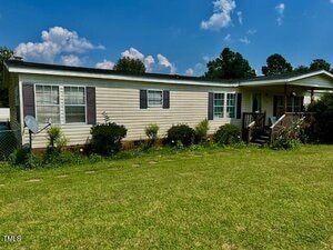 Located at 174 Daffodil Drive in Henderson, this single-story manufactured home features beige siding, dark shutters, a covered front porch, several windows, a satellite dish, and bushes along the foundation in a grassy yard.