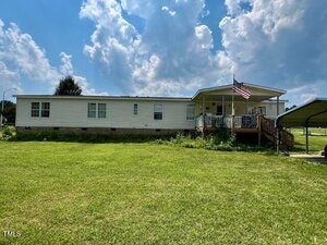 A single-story manufactured home with white siding, a covered front porch with an American flag, sits on a large grassy lawn. A metal carport is visible on the right under a partly cloudy sky.