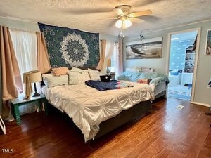 A bedroom with wood floors features a large bed with white bedding, decorative pillows, and a green side table. There is a tapestry on the wall, a ceiling fan, and a smaller bed near a doorway leading to a bathroom.