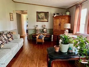 A living room with wood flooring, a sofa with patterned pillows, a wooden armoire, a coffee table with various potted plants, a wicker chair, wall art, and sunlight coming through curtained windows.