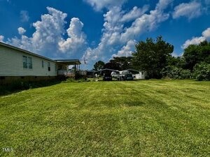 A large, neatly mowed grassy yard leads to a white manufactured home on the left, with several cars parked under metal carports in the background, and a partly cloudy blue sky overhead.