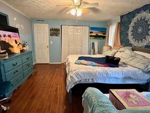 A bedroom with wooden floors, a large bed with white bedding, a blue dresser with a TV and decor, a tapestry on the wall, two closets with white doors, and a ceiling fan with lights.