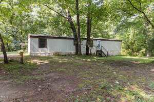 A white mobile home at 3170 Loblolly Lane sits in a wooded area of Wake Forest, with grass and trees surrounding it; the ground is patchy with dirt and leaves and a small set of stairs leads to the front door.