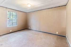 An empty room with beige walls, beige carpeting, a window with sunlight coming through, a ceiling light, and black trim along the baseboards and ceiling. Electrical outlets are visible on the walls.