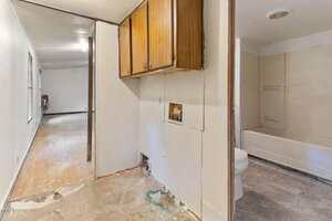 A hallway with worn floors leads to a room in the distance. An open area shows laundry hookups below wooden cabinets and a view into a bathroom with a toilet and bathtub. Surfaces appear unfinished or in need of repair.