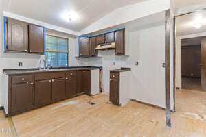 A partially renovated kitchen with brown cabinets, a black countertop, an empty space for an appliance, exposed outlets, and unfinished flooring. A doorway leads to another room with visible construction materials.