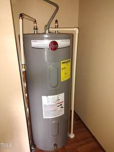 A gray Rheem water heater stands in a corner, connected to white and copper pipes. An "Energy Guide" label and instruction sticker are visible on the front. The unit is on a wooden floor against beige walls.