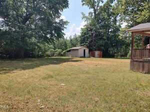 A grassy backyard with a few large trees, a small white shed, a red storage structure, and a wooden deck attached to a house on the right side. The sky is mostly clear with some clouds.