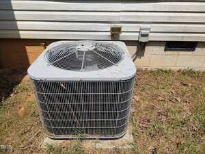 A large outdoor air conditioning unit sits on a concrete pad next to the exterior wall of a house, with electrical outlets and a vent visible on the wall behind it. Grass and dirt cover the ground around the unit.