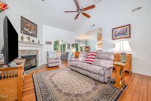 A living room with a beige couch, patterned rug, TV on a wooden stand, fireplace with mantel, armchair, hardwood floor, and ceiling fan. The space connects to a dining area through an arched doorway.