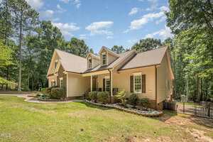A beige two-story house with stone accents, gabled roof, covered front porch, and white railings, surrounded by trees and landscaped with bushes and small rocks under a blue sky with scattered clouds.