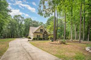 A beige, single-story house with a gabled roof sits among tall trees at 1513 Anterra Drive in Wake Forest. A wide concrete driveway curves to the entrance, with landscaping, potted plants near the front, and a partly cloudy sky above.
