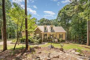 A two-story house with a stone and beige exterior sits among tall trees. The house has three dormer windows, a covered porch, and landscaped gardens in front. Sunlight filters through the surrounding forest.