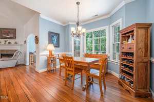 A dining room with wood flooring, a rectangular table and four chairs, a wooden cabinet with wine storage, large windows, a chandelier, and an adjacent living room visible through an arched doorway.