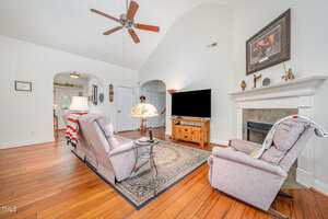 A living room with wood flooring, two recliners facing a TV on a wooden stand, a rug, a fireplace with decor above it, a ceiling fan, and a lamp on a side table. An arched entryway leads to another room.