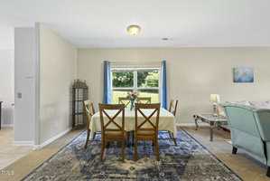 A dining area with a rectangular table covered by a cream tablecloth, four wooden chairs, a blue patterned rug, a vase with flowers, and a window with blue curtains overlooking greenery. Part of a living room is visible.