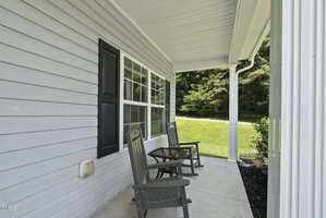 A covered front porch with two black rocking chairs on a concrete floor, white siding, black shutters, and a view of a grassy yard with trees in the background.