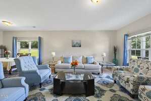 A living room with a beige sofa, two blue armchairs, a floral armchair, a glass coffee table with a vase of flowers, blue curtains, and large windows letting in natural light. A dining area is visible in the background.