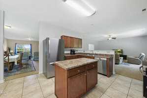 A spacious open-concept kitchen with brown cabinets, an island, stainless steel appliances, and tile flooring, leading to a carpeted living area and a dining room with a table and chairs in the background.