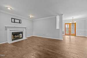 A spacious, empty living and dining area with light gray walls, hardwood floors, a white fireplace, ceiling lights, and a chandelier over the dining space near glass doors leading outside.