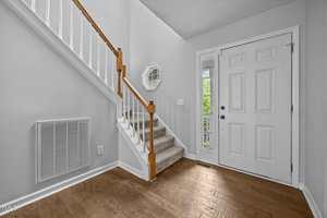 A foyer with hardwood flooring, a white front door, a carpeted staircase with wooden handrail, a wall vent, light gray walls, a white trim, and a hexagonal mirror above the light switch.