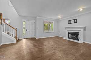 A living room with hardwood floors, a white fireplace, a window, and a staircase with white railings. The walls are painted light gray and there are recessed ceiling lights. The front door is near the stairs.