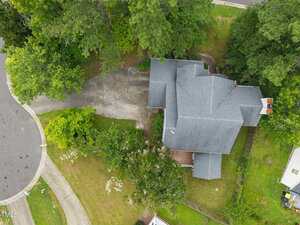 Aerial view of a house with a gray roof, surrounded by trees and greenery. The property features a paved driveway, a wooden fence, and is located near a cul-de-sac with a curved sidewalk.