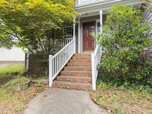 A set of brick steps with white railings leads up to a red front door on a brick house, surrounded by green bushes and a tree. The pathway and yard show some overgrown plants and leaves.