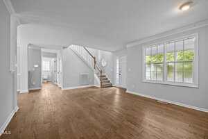 Unfurnished living room with hardwood floors, white walls, large front window, and a staircase leading upstairs. A hallway to the left leads to another room and a bathroom. Ceiling lights are on, and natural light enters the room.