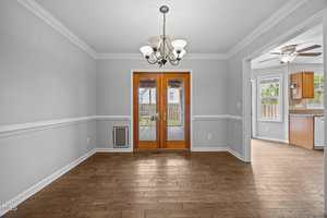 A bright dining room with light gray walls, white trim, wood flooring, a ceiling chandelier, and double glass doors leading to a deck. A kitchen with wood cabinets is visible through an open doorway.