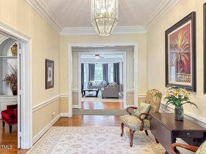A hallway with light yellow walls and wood flooring features a rug, a chair with a cushion, a console table with a vase of flowers, framed art, and a chandelier. The hallway leads to a bright living room.