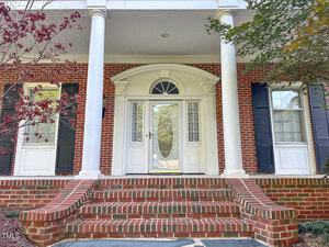A brick front porch with curved steps leads to a white door with decorative glass flanked by sidelights and large white columns. The entry is framed by red brick, black shutters, and white trim.