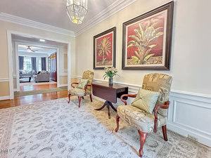 A hallway with two patterned armchairs, a small dark wood table with a plant, two large framed palm artwork pieces on the wall, and a light-colored rug; living room visible in the background.