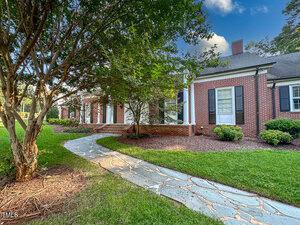A flagstone walkway curves through a neatly landscaped yard toward a red brick house with white trim and black shutters. A large tree partially shades the walkway and the house's front entrance.