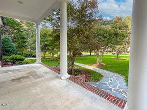 A covered porch with white columns overlooks a landscaped yard with a stone walkway curving through green grass and trees. The porch has a concrete floor and red brick trim along the edge.