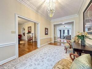 A hallway with cream walls, white trim, and crown molding features a light fixture, a white rug, a bench with cushions, and a table with flowers. Doorways lead to a living room and another furnished room.