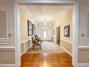 A bright entry hallway with hardwood floors, beige walls, white trim, a patterned rug, chairs with a small table, artwork on the walls, and a glass front door with sidelights and a decorative light fixture.