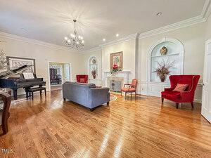 A spacious living room with hardwood floors, a grand piano, a gray sofa, two red armchairs, built-in shelves, a fireplace, floral arrangements, and framed artwork on the wall.