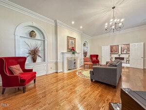 A living room with hardwood floors, a gray sofa, two red armchairs, a fireplace with framed art above it, built-in shelves with vases, a chandelier, and double doors leading to another room.