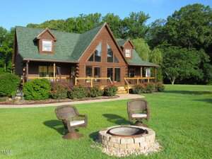 A log cabin with large windows and a green roof sits behind a neatly trimmed lawn. Three wicker chairs are arranged around a stone fire pit in the foreground, and trees and bushes surround the house.