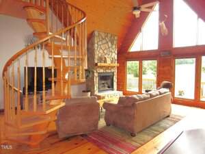A living room with a spiral wooden staircase, stone fireplace, two brown sofas, a TV, and large windows letting in natural light. The space features wood-paneled walls and ceiling, and overlooks an outdoor area.