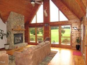 A living room with wood-paneled walls and ceiling, a stone fireplace, large windows overlooking a green yard, brown couches, a small coffee table, and a mounted deer head above the windows.