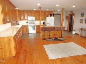 Spacious kitchen with wooden cabinets and flooring, a large countertop, stainless steel stove and oven, three bar stools at an island, and a light-colored rug on the floor.