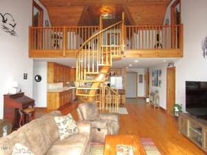 Open living room features wood floors, a spiral staircase leading to a loft with railing, vaulted wood ceiling, kitchen with wood cabinets, a couch and coffee table, piano, wall-mounted TV, and framed pictures on the walls.