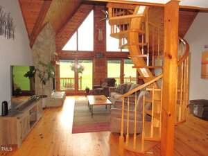 A living room with wood-paneled walls, large windows, a spiral wooden staircase, a stone fireplace, and plush seating. The floor is wooden and a TV is positioned on a stand in the corner.