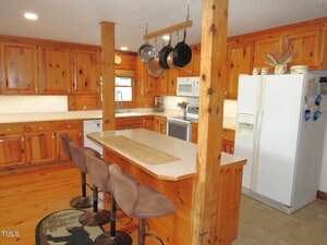 A kitchen with wooden cabinets and paneling, a white refrigerator, a built-in microwave, three brown barstools at a counter, a hanging pot rack, and a small rug on the wood floor.