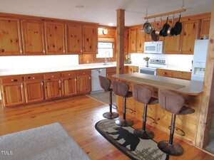 Kitchen with wooden cabinets and floors, white countertops, stainless steel sink, stove, microwave, and a counter with four brown barstools. Pots and pans hang above the counter, and a bear-patterned rug is on the floor.