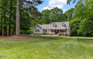 A light gray, two-story house at 450 S Stratford Drive, Henderson, features a covered front porch and a gabled roof. It sits on a large grassy lawn surrounded by tall trees, with a black car parked in the driveway under a partly cloudy sky.